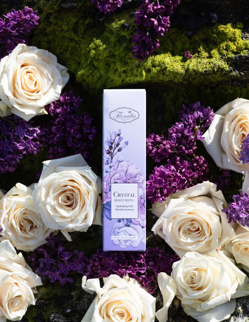 Floralla Crystal Moisturizing Essence packaging with hydrating anti-wrinkle formula, displayed among white roses and purple lilacs on a green moss background, representing natural skincare and beauty.
